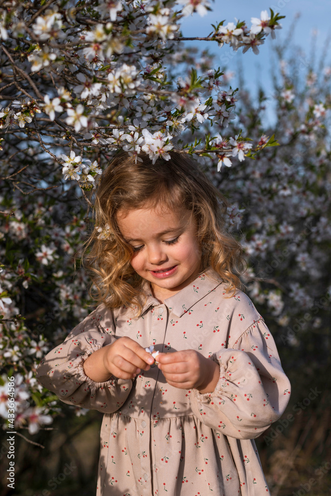Adorable smiling little child in dress standing near blossoming tree with flowers in spring park and looking down Adorable smiling little child in dress standing near blossoming tree with flowers in spring park and looking down