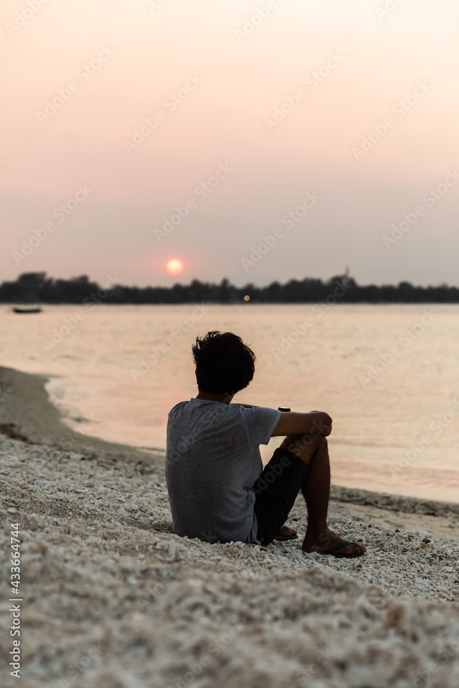 Back view full length unrecognizable male in casual summer wear resting on sandy seashore and enjoying picturesque sunset view Back view full length unrecognizable male in casual summer wear resting on sandy seashore and enjoying picturesque sunset view