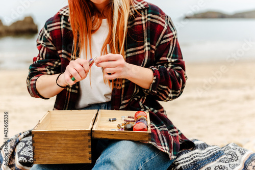 Crop anonymous craftswoman creating handmade bijouterie while using semiprecious stones and sitting on beach near sea photo