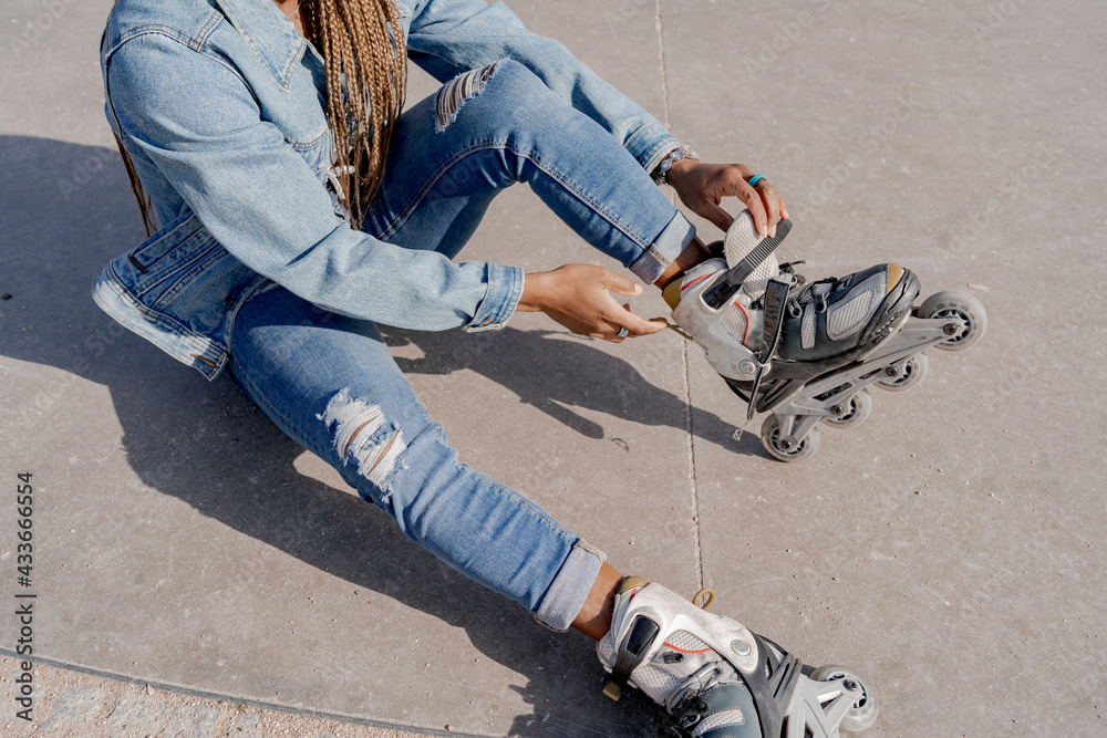 From above of African American female in denim outfit putting on rollerblades on sunny day in skate park From above of African American female in denim outfit putting on rollerblades on sunny day in skate park