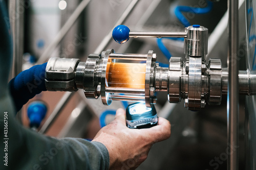 Crop unrecognizable worker with light under alcoholic liquid with grain particles passing through filter tube of metal equipment in brewery