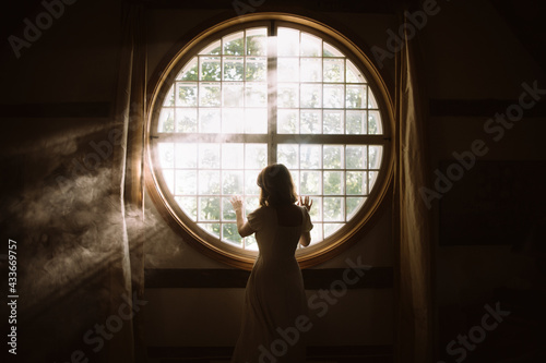 Back view of unrecognizable gentle female touching fence on round shaped window in house in sunlight photo