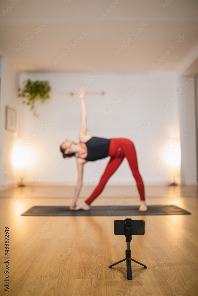 High angle side view of slim female in Trikonasana pose and shooting live video on smartphone during yoga training High angle side view of slim female in Trikonasana pose and shooting live video on smartphone during yoga training