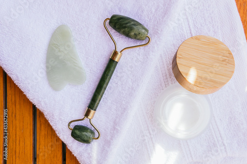 High angle of jade roller and guasha scraper placed on wooden table with towel and natural facial cream in spa salon