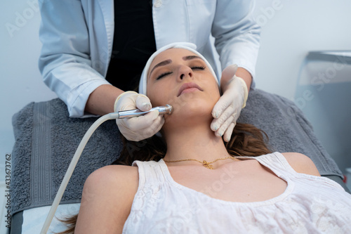 Cropped unrecognizable professional cosmetologist using special equipment and doing microdermabrasion facial treatment for female client in modern beauty clinic photo