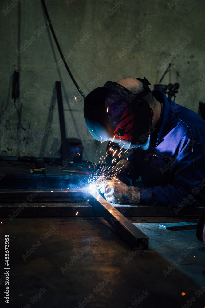 Crop faceless worker in gloves and uniform welding metal details on table near constructions in factory Crop faceless worker in gloves and uniform welding metal details on table near constructions in factory