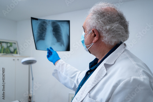 Side view of elderly male doctor in mask and gloves examining x ray image of lungs of patient during coronavirus in hospital photo