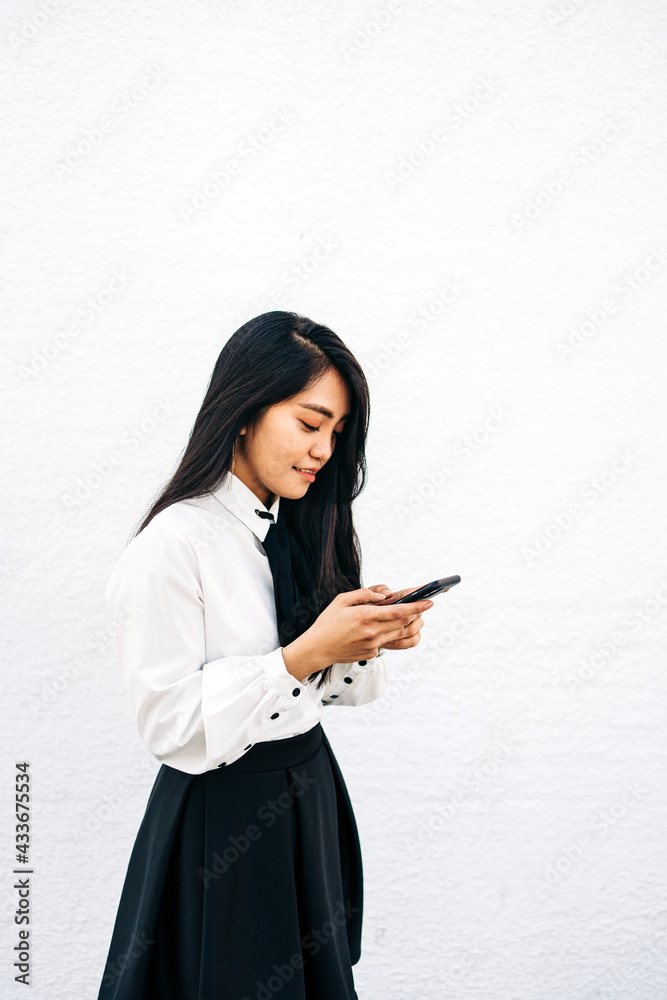 Side view of teen ethnic female in formal outfit standing near white wall and using mobile phone