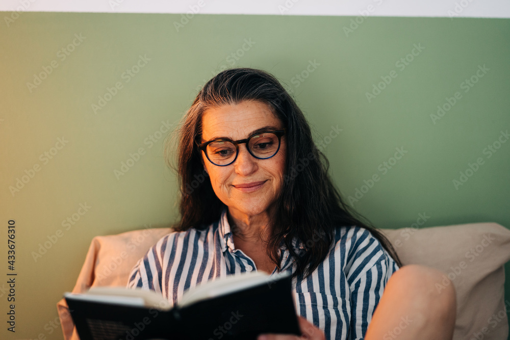 Relaxed mature Hispanic woman in casual clothes and eyeglasses sitting in bed and reading interesting book before sleeping Relaxed mature Hispanic woman in casual clothes and eyeglasses sitting in bed and reading interesting book before sleeping