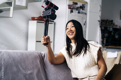 Smiling ethnic female vlogger recording video on photo camera while sitting on couch in living room photo
