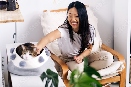 Smiling ethnic female sitting with crossed legs in armchair and caressing charming cat in house room photo