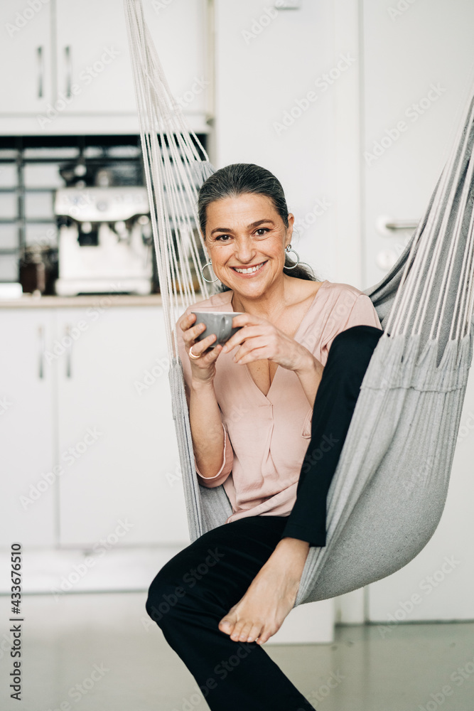 Positive middle aged female drinking hot beverage while sitting on hammock in kitchen with modern kitchenware looking at camera on blurred background