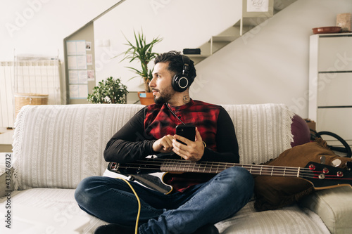 Adult bearded male musician with bass guitar in headphones text messaging on cellphone in living room