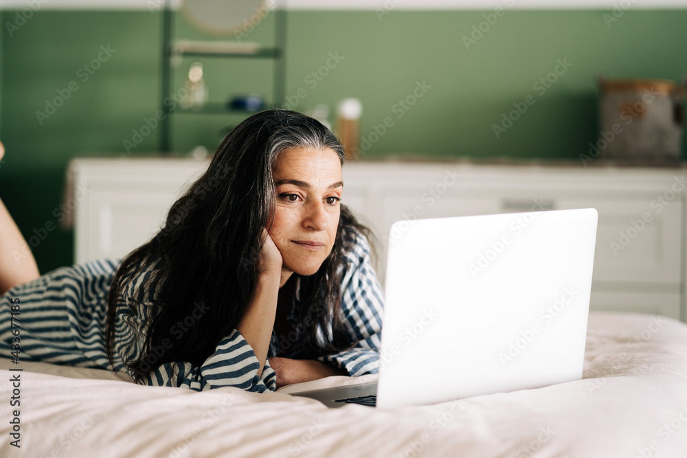 Side view of focused mature self employed Hispanic woman with long dark hair in casual clothes lying on bed with laptop during online work at home Side view of focused mature self employed Hispanic woman with long dark hair in casual clothes lying on bed with laptop during online work at home