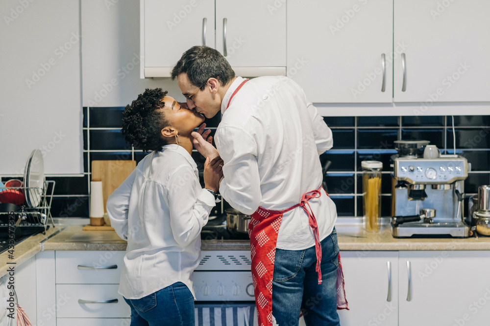 Side view of multiethnic couple in love kissing while standing in kitchen and cooking Side view of multiethnic couple in love kissing while standing in kitchen and cooking