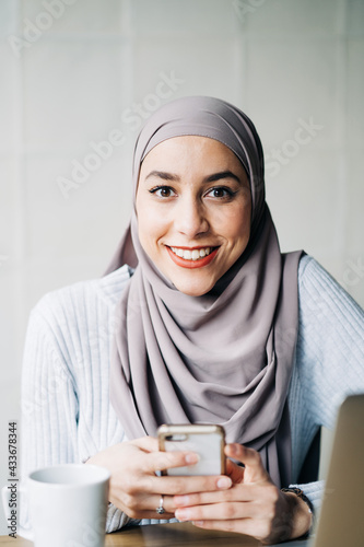 Portrait of ethnic female freelancer in hijab using smartphone while sitting at table in cafe and working remotely