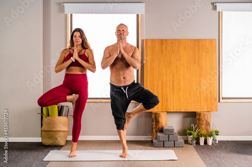 Full body of concentrated couple in sportswear performing Vrikshasana pose while practicing yoga in studio with light interior photo