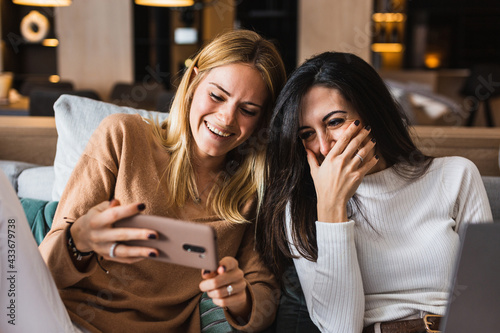 Couple of homosexual females sitting on sofa and watching funny video on mobile phone while laughing and having fun