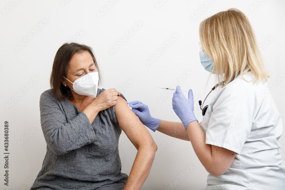 Female medical specialist in protective uniform and latex gloves vaccinating senior female patient in clinic during coronavirus outbreak Female medical specialist in protective uniform and latex gloves vaccinating senior female patient in clinic during coronavirus outbreak