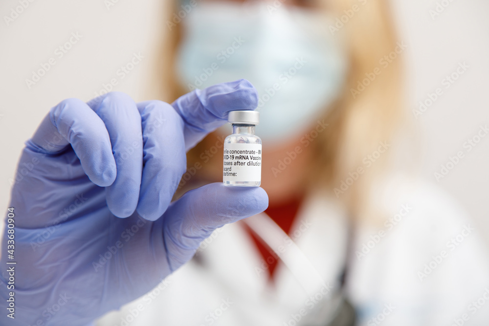 Female medic in protective face shield mask and latex gloves with vial of coronavirus vaccine showing to camera while standing in hospital room Female medic in protective face shield mask and latex gloves with vial of coronavirus vaccine showing to camera while standing in hospital room
