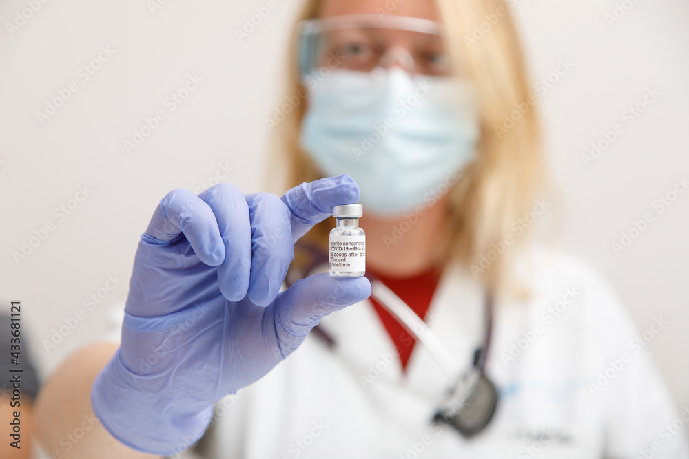 Female medic in protective face shield mask and latex gloves with vial of coronavirus vaccine showing to camera while standing in hospital room Female medic in protective face shield mask and latex gloves with vial of coronavirus vaccine showing to camera while standing in hospital room