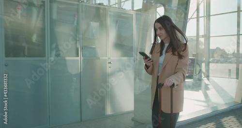 Stylish business woman walking with suitcase near airport and using modern smartphone. Successful female entrepreneur solving working problems before trip.