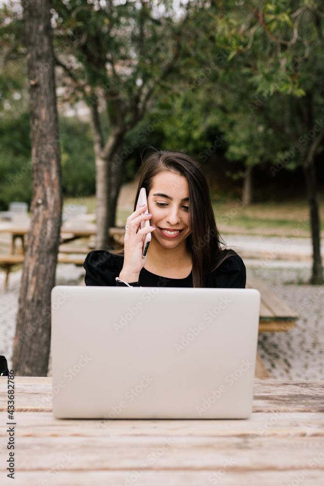 Female entrepreneur sitting at table with laptop in park and speaking on smartphone while working remotely Female entrepreneur sitting at table with laptop in park and speaking on smartphone while working remotely