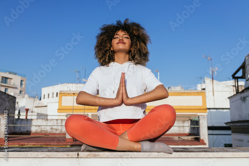 Tranquil African American female sitting in Padmasana with Namaste gesture and meditating with closed eyes while practicing yoga on rooftop