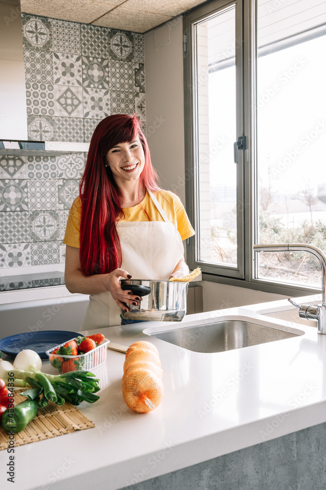 Young homosexual female pouring water from tap into saucepan with uncooked pasta against assorted vegetables in house Young homosexual female pouring water from tap into saucepan with uncooked pasta against assorted vegetables in house