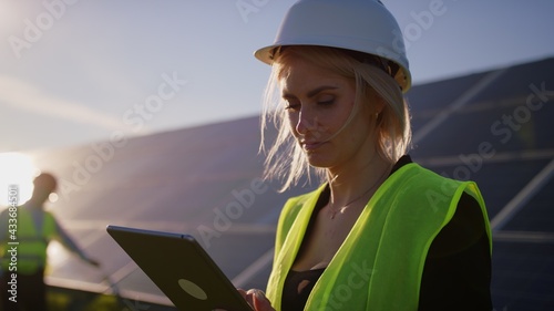 Portrait of a female solar engineer standing uses a tablet and looking at the camera, another man near the panels and entering information into a laptop. Green energy concept