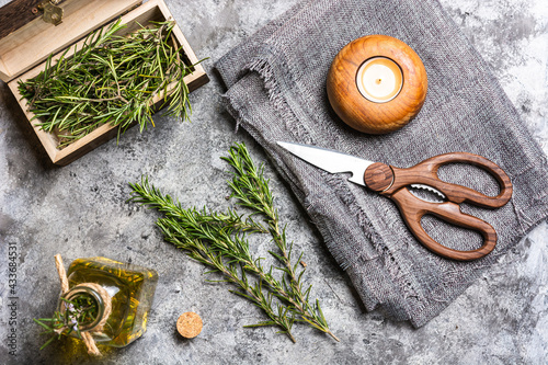 Top view of rosemary sprigs with green leaves in small chest near glass bottle with oil and scissors near candle in candlestick on textile on table