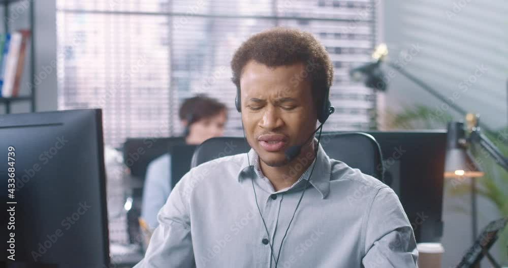 Close up portrait of young handsome African American male operator speaking in headset videochatting on computer sitting at desk in office Man employee talking with client on online call work concept Close up portrait of young handsome African American male operator speaking in headset videochatting on computer sitting at desk in office Man employee talking with client on online call work concept