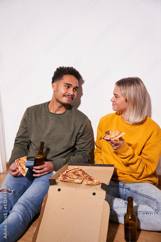 Cheerful multiethnic couple sitting on floor at home while eating delicious pizza and drinking beer together Cheerful multiethnic couple sitting on floor at home while eating delicious pizza and drinking beer together