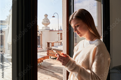 Side view of calm young lady in stylish sweater messaging on smartphone while standing near window at home photo
