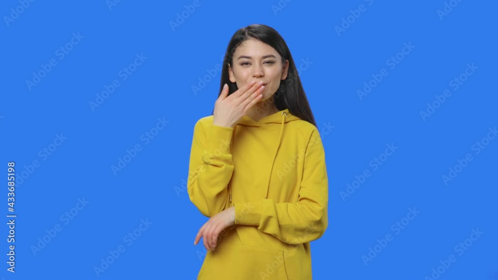 Portrait of brunette showing heart with fingers then blowing kiss. Young woman with long hair in yellow sweatshirt posing in studio with blue screen background. Close up. Slow motion ready 59.94fps. Portrait of brunette showing heart with fingers then blowing kiss. Young woman with long hair in yellow sweatshirt posing in studio with blue screen background. Close up. Slow motion ready 59.94fps.