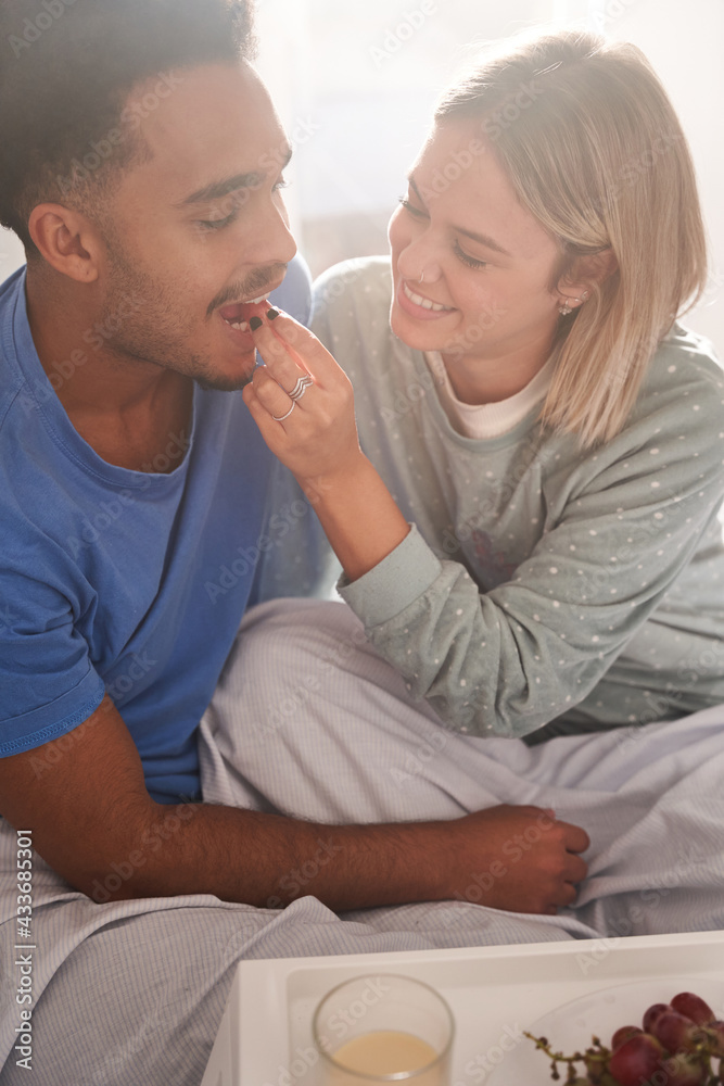 High angle of multiethnic couple in love feeding each other while sitting on bed and having tasty breakfast in morning High angle of multiethnic couple in love feeding each other while sitting on bed and having tasty breakfast in morning