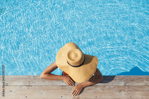 Enjoying summer vacation. Stylish woman in hat relaxing in pool water at wooden pier, top view. Slim young female sunbathing at  swimming pool edge. Travel and Holidays. Space for text photo