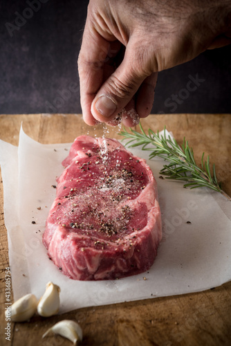 Crop anonymous male cook seasoning uncooked meat piece with salt and ground black pepper between garlic cloves and fresh rosemary sprig