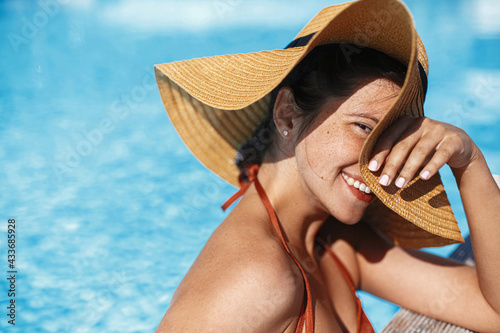 Beautiful happy carefree woman in hat relaxing in pool, enjoying summer vacation. Portrait of smiling young female sunbathing and resting at swimming pool at tropical resort. Holidays and travel