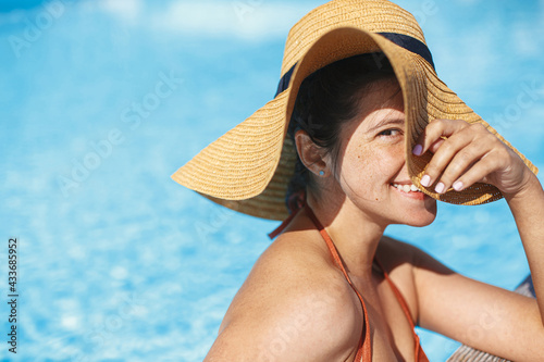 Beautiful happy carefree woman in hat relaxing in pool, enjoying summer vacation. Portrait of smiling young female sunbathing and resting at swimming pool at tropical resort. Holidays and travel