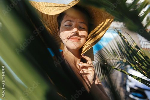 Obraz na plátně Portrait of beautiful carefree woman in hat relaxing under palm leaves at tropical resort, enjoying summer vacation