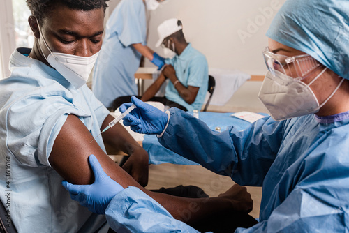 Female doctor in protective uniform and latex gloves vaccinating male African American patient in clinic during coronavirus outbreak photo