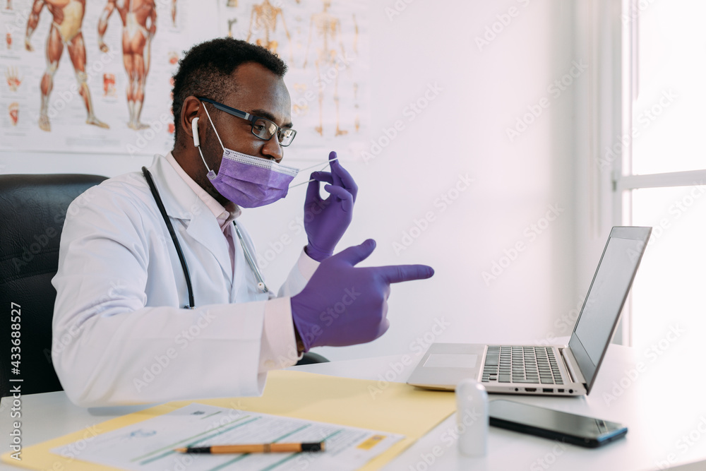 African American doctor in eyeglasses and face mask working with online patient on netbook while writing in patient file at table in hospital