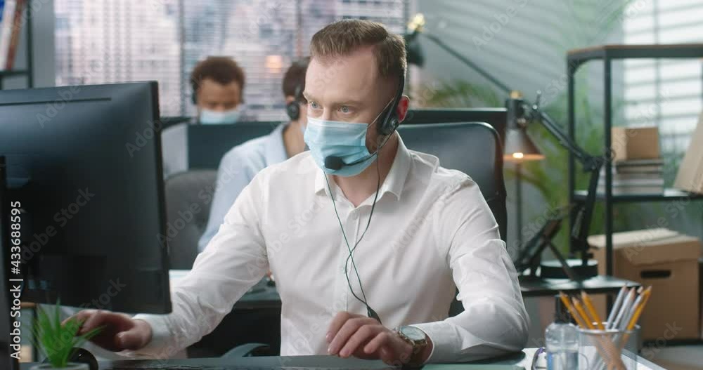 Portrait of young handsome Caucasian male worker in medical mask sitting at table in cabinet in headset, typing on computer browsing online. Busy man manager at work in quarantine, job concept Portrait of young handsome Caucasian male worker in medical mask sitting at table in cabinet in headset, typing on computer browsing online. Busy man manager at work in quarantine, job concept