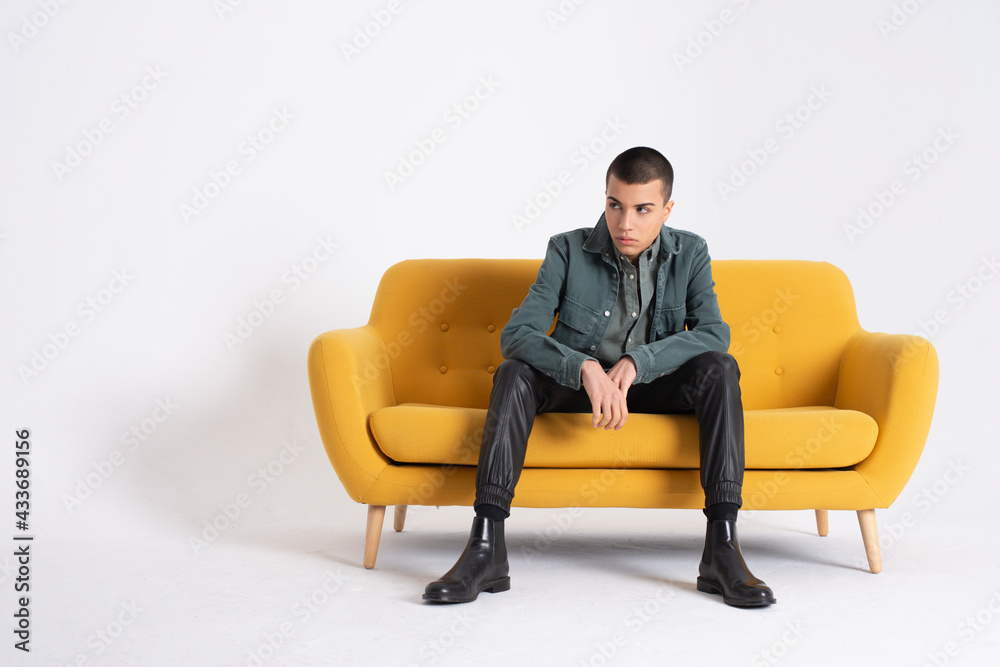 Young handsome male model in trendy clothes sitting on yellow couch on white background in studio and looking away Young handsome male model in trendy clothes sitting on yellow couch on white background in studio and looking away