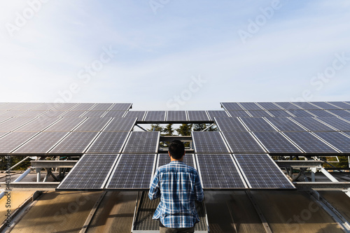 Back view anonymous professional technician wearing checkered shirt checking photovoltaic panels in solar power station on clear sunny weather