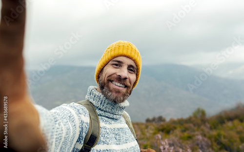 Cheerful bearded male explorer with backpack and in warm clothes taking selfie in mountains and smiling at camera