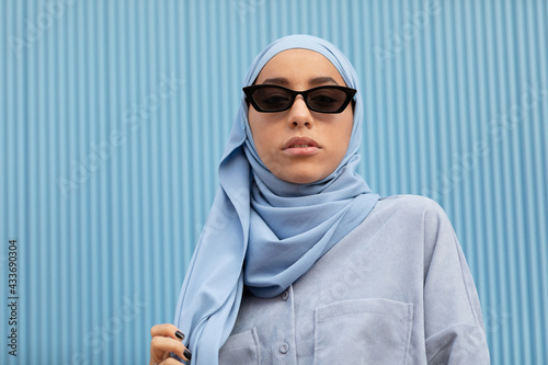 Young contemplative ethnic female in blue headscarf and modern sunglasses looking at camera in daylight