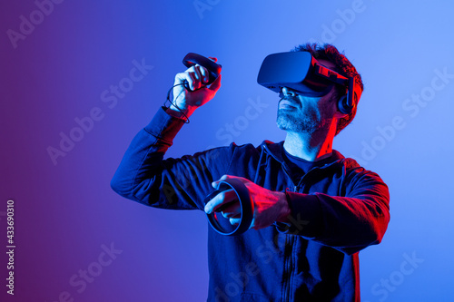 Anonymous unshaven male in hoodie and modern goggles with controllers and extended arm experiencing virtual reality photo