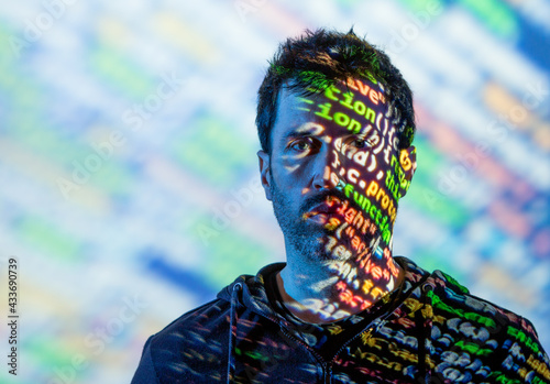 Adult bearded male in hoodie with multicolored words on half of face from projector light looking at camera photo